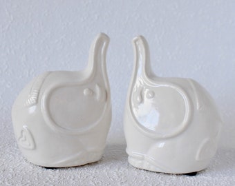 May include: Two white ceramic elephant figurines with heart details. The elephants are facing each other and have a glossy finish.