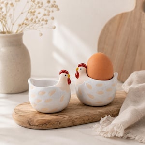 May include: Two white ceramic egg cups shaped like chickens with red combs and yellow beaks, featuring light tan spots. One egg cup is empty, while the other holds a single brown egg. The chicken-shaped holders have a smooth, glossy finish.