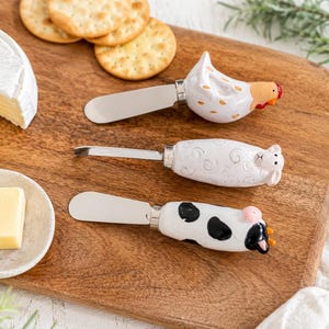 May include: Three cheese knives with ceramic handles shaped like a chicken, a sheep, and a cow. Each knife features a stainless steel blade and a detailed, hand-painted animal figure. The chicken is white with orange accents, the sheep is white with textured wool, and the cow is white with black spots and pink ears.