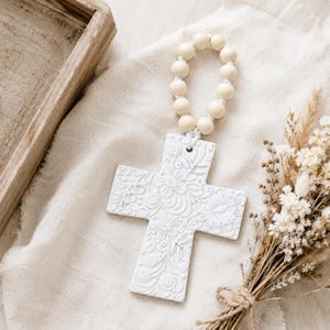 Puede incluir: Un adorno de cruz de cerámica blanca con un patrón floral en relieve en su superficie. La cruz está unida a un lazo de cuentas de madera redondas y claras, creando una pieza decorativa colgante con un acabado mate y texturizado.