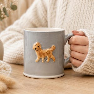 Könnte beinhalten: Graue Keramiktasse mit einem geformten goldenen Hund auf der Seite. Die Tasse hat einen abgerundeten Griff und einen weißen Innenraum. Die Tasse steht auf einer Holzoberfläche.