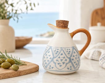 Może przedstawiać: Ceramiczna oliwiarka z białą podstawą, ozdobiona jasnoniebieskim geometrycznym wzorem na korpusie. Naczynie posiada zakrzywiony brązowy uchwyt, szeroki dzióbek oraz cylindryczny korek, który szczelnie zamyka otwór w górnej części naczynia.
