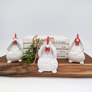 May include: Three white ceramic chickens with red combs and wattles, each covering their eyes with their wings. The chickens are sitting on a wooden surface in front of a stack of books with the text "welcome to our home" printed on the top book.