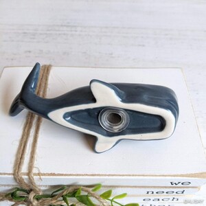 May include: A ceramic whale hook in a dark blue glaze with a white underbelly. The hook is designed to be mounted on a wall and has a hole in the back for hanging.