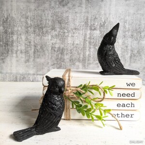 May include: A stack of white books with black text that reads "we need each other". Two black bird figurines are placed on top of the books. The books are tied together with twine and a sprig of greenery.