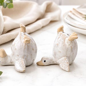 May include: Two ceramic duck figurines in a glossy white glaze with tan accents on the beaks, feet, and wings. Each duck is depicted in a playful pose with its head lowered and tail pointed upward, featuring small black eyes and textured feathers.