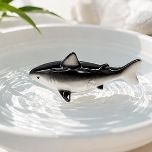 Può includere: Una piccola statuetta di squalo in ceramica lucida con dorso nero e ventre bianco, che galleggia in una ciotola bianca circolare poco profonda piena d'acqua limpida, creando lievi increspature concentriche sulla superficie attorno al corpo.