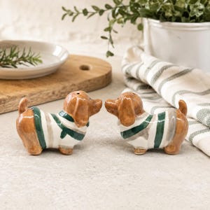 May include: Two ceramic dachshund salt and pepper shakers, each featuring a glossy brown finish and wearing a white sweater with horizontal dark green and light gray stripes. The two dog-shaped shakers are positioned facing each other.