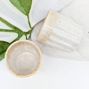 May include: Two small, handmade ceramic cups. One is upright, the other is tilted, revealing a textured exterior and speckled interior. The cups have a light beige color with darker brown rims. The background includes a white cloth and a green and white leaf.