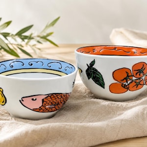 Puede incluir: Dos cuencos de cerámica blanca con ilustraciones pintadas a mano. El cuenco izquierdo muestra un pez rosa y un limón amarillo con un borde azul. El cuenco derecho presenta un racimo de tomates naranjas y hojas de albahaca verde con un borde naranja.