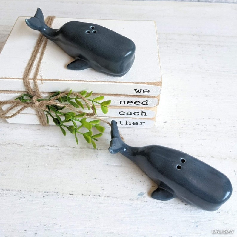 May include: A set of two gray whale salt and pepper shakers. The shakers are sitting on a stack of books with the words "we need each other" written on them.