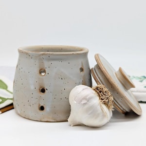May include: A ceramic garlic keeper with a lid, featuring a speckled glaze in shades of grey and brown. The keeper has holes for air circulation and is shown with a whole garlic bulb.