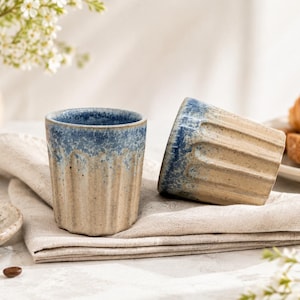 May include: Two ceramic cups with a fluted, ribbed exterior design. The cups feature a speckled beige base colour that transitions into a textured, mottled blue glaze around the upper rim. The cups have a cylindrical shape with vertical grooves.