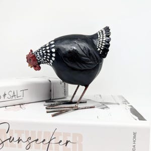 Puede incluir: Figurita de pollo de cerámica negra y blanca con detalles rojos. El pollo está de pie sobre patas metálicas delgadas y tiene un patrón de manchas en su cuerpo.