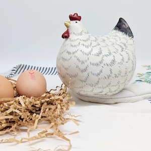 Könnte beinhalten: Keramik-Hühnerfigur mit weißem Körper, schwarzen Markierungen und rotem Kamm und Kehllappen. Das Huhn befindet sich neben einem Nest aus braunem geschreddertem Papier mit zwei braunen Eiern. Der Hintergrund ist weiß.