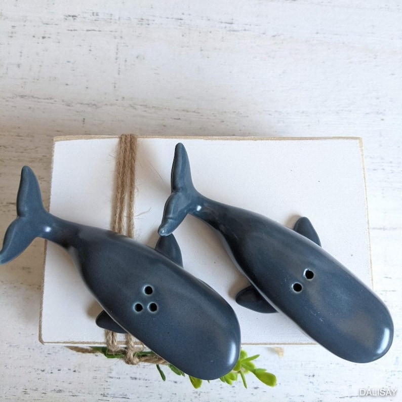 May include: Two blue ceramic whale salt and pepper shakers on a white wooden surface. The whales are facing each other with their tails in the air.