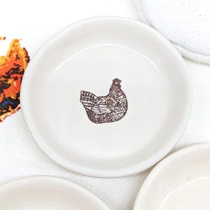 Puede incluir: Pequeño plato de cerámica blanquecina con una ilustración marrón de una gallina en el centro. El plato tiene forma redondeada y un borde ligeramente elevado. La ilustración de la gallina es detallada.