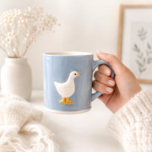May include: A light blue ceramic mug with a white goose design. The mug has a handle and a white interior. The goose has yellow feet and a yellow beak. The mug is held by a person wearing a white sweater.