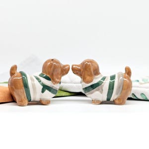 May include: A pair of ceramic dachshund salt and pepper shakers. The shakers are brown with white and green striped sweaters. They are facing each other.