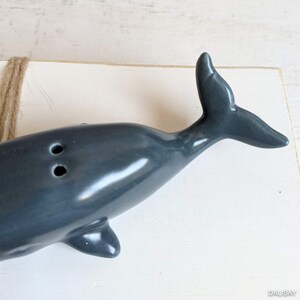 May include: A gray ceramic whale salt and pepper shaker set. The whale is in a horizontal position with its tail raised. The whale has two holes on its back for dispensing salt and pepper.