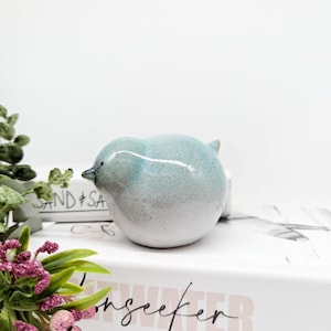 Puede incluir: Una pequeña figurita de pájaro de cerámica en un esmalte azul claro y blanco. El pájaro está sentado en un libro blanco con la palabra "Seawater" impresa en la portada.