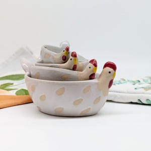 May include: Set of four white ceramic measuring cups shaped like chickens with red and yellow accents. The cups are nested inside each other.