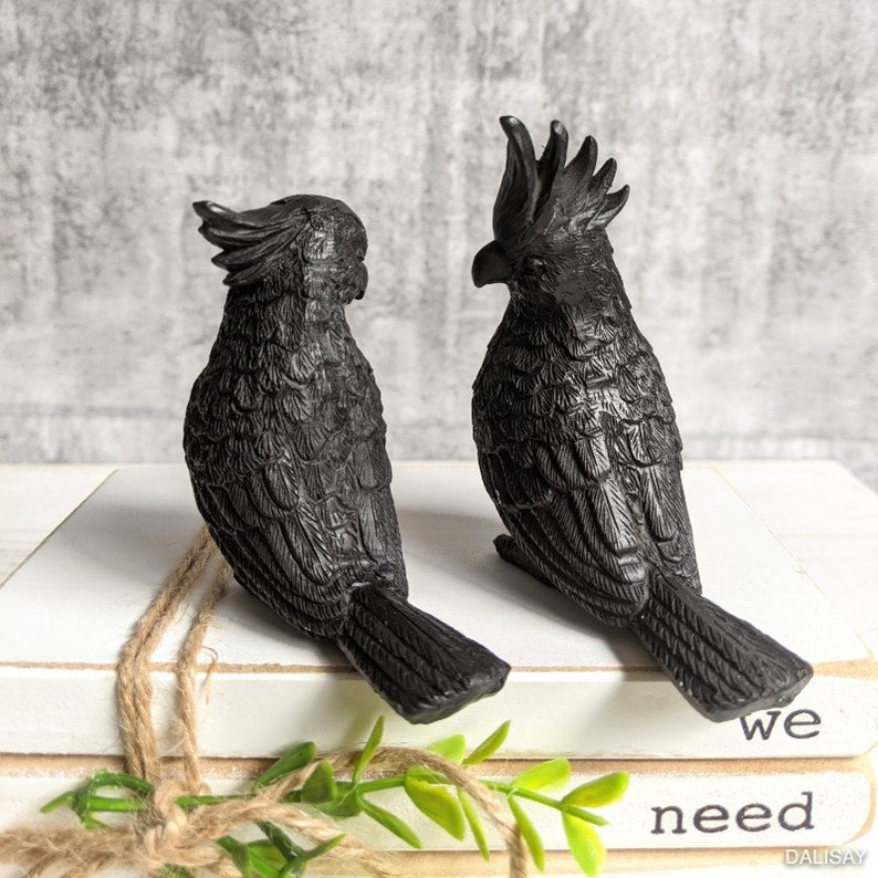May include: Two black bird figurines with detailed feather patterns. The birds are perched on a stack of books with the words "we need" visible on the top book.