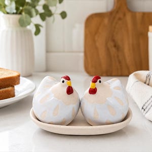 Puede incluir: Un par de saleros y pimenteros de cerámica con forma de gallina en tonos azul claro y crema, con crestas rojas y picos amarillos, colocados juntos sobre una pequeña bandeja redonda de cerámica color crema en una encimera.