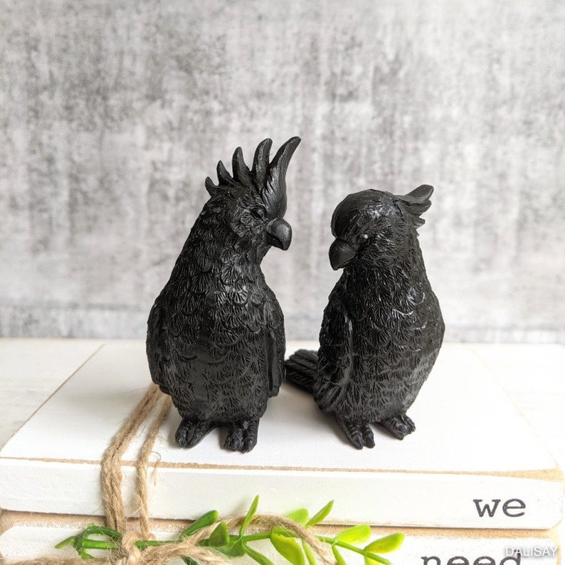 May include: Two black, feathered, bird figurines. The birds are perched on a stack of white books with the word "we" visible on the top book.