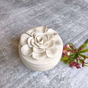 May include: A white ceramic trinket box with a raised flower design on the lid. The flower is made up of many petals and has a delicate, feminine look.