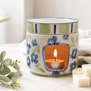 Puede incluir: Un calentador de cera de cerámica con un patrón floral azul pintado a mano sobre una base blanca. El plato superior tiene rayas verdes y azules. Una vela de té encendida se encuentra en la abertura arqueada, junto a tres cubos de cera blancos.