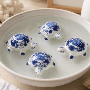 May include: Four small ceramic turtle figurines floating in a shallow, round, off-white bowl filled with water. Each turtle features a white base with a detailed blue floral pattern on its shell, head, and flippers, creating a cohesive set.