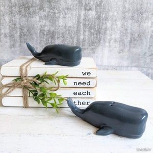 May include: Two gray whale salt and pepper shakers on a stack of white books with the text "we need each other" written on the books.