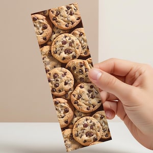 May include: A rectangular bookmark featuring a pattern of chocolate chip cookies. The cookies are golden brown with dark chocolate chips. The bookmark is held by a hand against a neutral background.