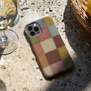This phone case features a soft, muted checkerboard pattern made up of square blocks in earthy tones—dusty pink, cream, sage green, mustard yellow, brown, and light blue. Each square has a slightly textured, vintage look, creating a warm, cosy, quilt-like aesthetic. No additional graphics or text appear on the case the buyer receives.