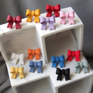 May include: Assortment of colorful bow-shaped hair clips in various colors, including red, pink, purple, blue, yellow, orange, white, and black. The clips are arranged on a white shelf with multiple compartments.