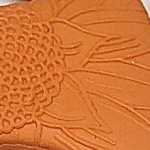 May include: Close-up of an orange clay relief sculpture of a sunflower. The centre of the sunflower is textured and the petals are raised.