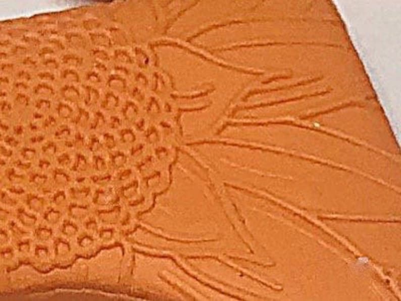 May include: Close-up of an orange clay relief sculpture of a sunflower. The centre of the sunflower is textured and the petals are raised.