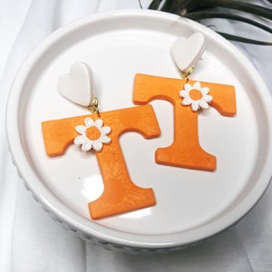 May include: A pair of orange letter T earrings with white flower accents and white heart-shaped tops.