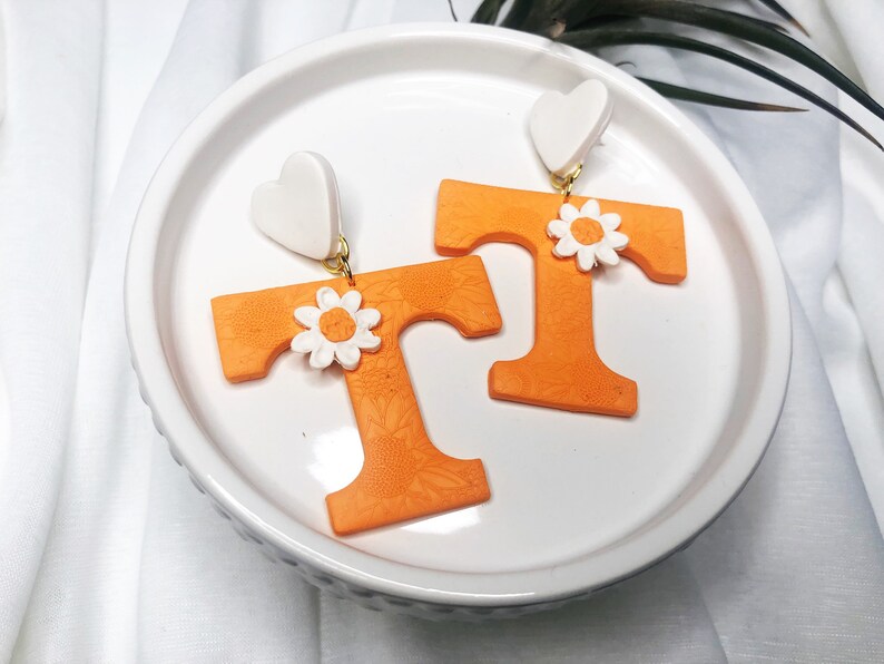 May include: A pair of orange letter T earrings with white flower accents and white heart-shaped tops.
