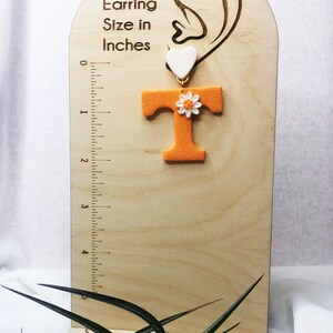 May include: Orange letter T with a white daisy and a white heart charm. The earring is shown on a wooden display with a ruler measuring up to 12.7 cm.