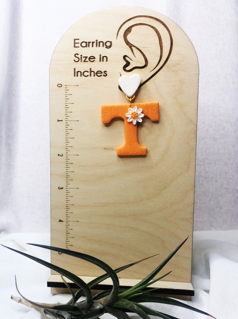 May include: Orange letter T with a white daisy and a white heart charm. The earring is shown on a wooden display with a ruler measuring up to 12.7 cm.