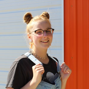 May include: A young person with blonde hair in two buns, wearing black framed glasses, a black t-shirt with a graphic design, and blue denim overalls. The person is smiling and standing in front of a white and orange wall.