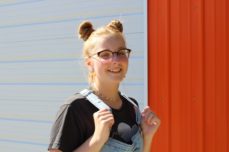 May include: A young person with blonde hair in two buns, wearing black framed glasses, a black t-shirt with a graphic design, and blue denim overalls. The person is smiling and standing in front of a white and orange wall.