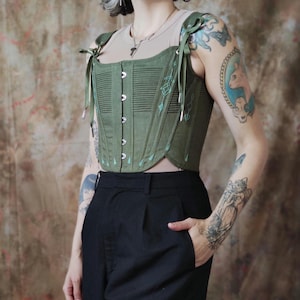 Könnte beinhalten: Ein grünes Korsett mit einem hellblauen Blumendesign und silbernen Ösen. Das Korsett hat verstellbare Träger und eine geschnürte Struktur. Das Model trägt eine schwarze Hose.