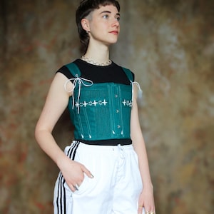 May include: A dark teal corset top featuring white star-shaped embroidery across the chest, white lace-up shoulder straps, and a front button closure. The garment is worn over a black short-sleeved shirt paired with white athletic pants with black stripes.