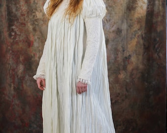 May include: A floor-length off-white dress featuring a high collar with a row of buttons, long sleeves with lace detailing, and a gathered bodice. The garment has a textured, crinkled fabric finish and a loose, flowing silhouette that extends to the floor.