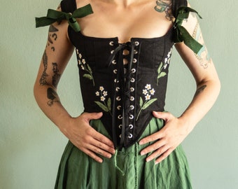May include: A black corset top featuring a front lace-up closure with silver-toned metal eyelets and black ribbon. The garment is decorated with embroidered white flowers and green leaves on both sides and includes dark green ribbon shoulder ties.