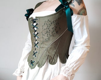 May include: An olive green corset featuring dark green ribbon lacing at the front and shoulders, with embroidered vine patterns on the side. The garment has a scalloped hem and is worn over a white long-sleeved blouse with lace-trimmed cuffs.