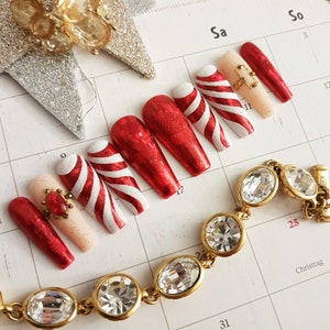Könnte beinhalten: Rote und weiße Zuckerstangen-Nagelspitzen mit Glitzer und ein einzelner Nagel mit einem roten Juwel. Die Nagelspitzen sind auf einem Kalender mit dem Wort "Weihnachten" darauf angeordnet.